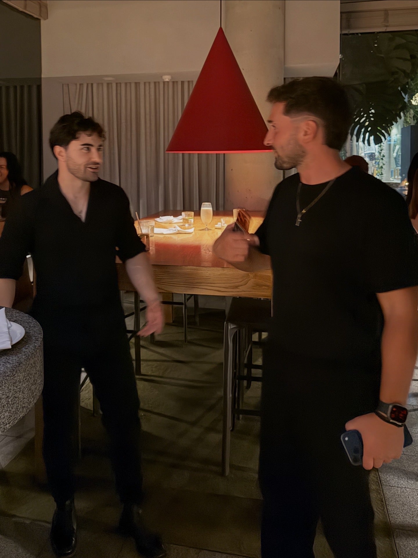 Two men in black shirts shaking hands in a restaurant setting with a red pendant light.