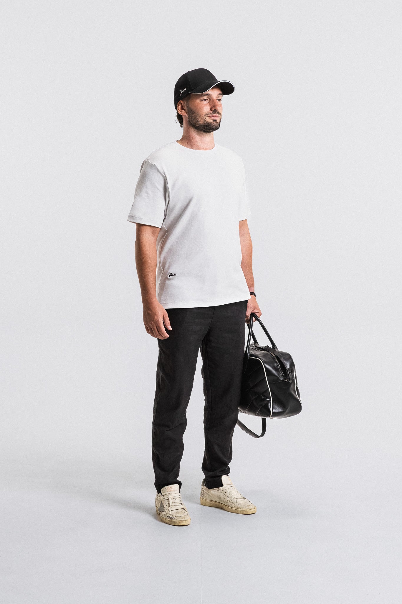 Man wearing a white t-shirt, black pants, and a black cap, holding a black bag on a light gray background