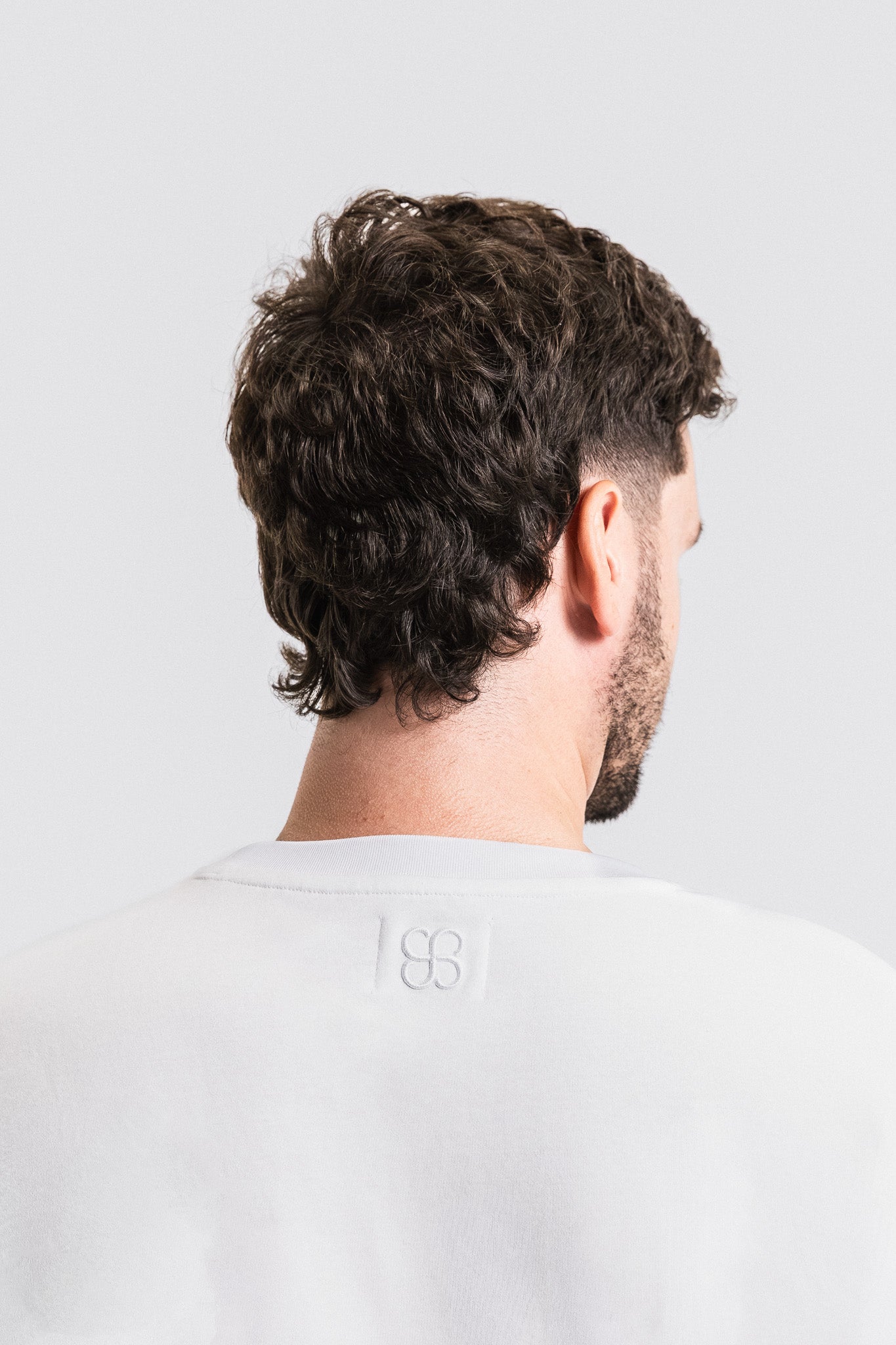 Man with wavy hair and a beard wearing a light gray t-shirt on a white background