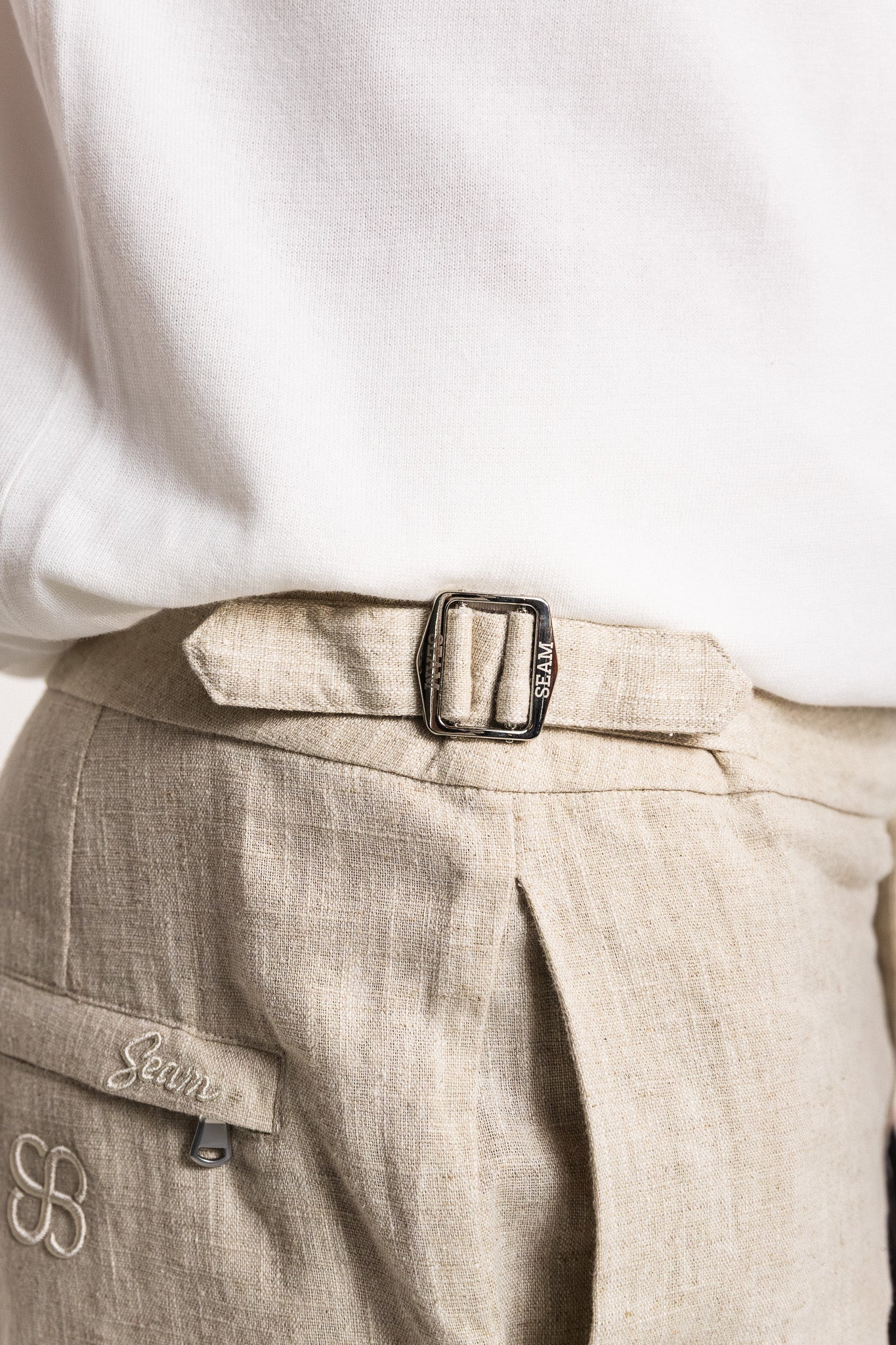 Close-up of beige pants with a belt and visible brand logo on a white background