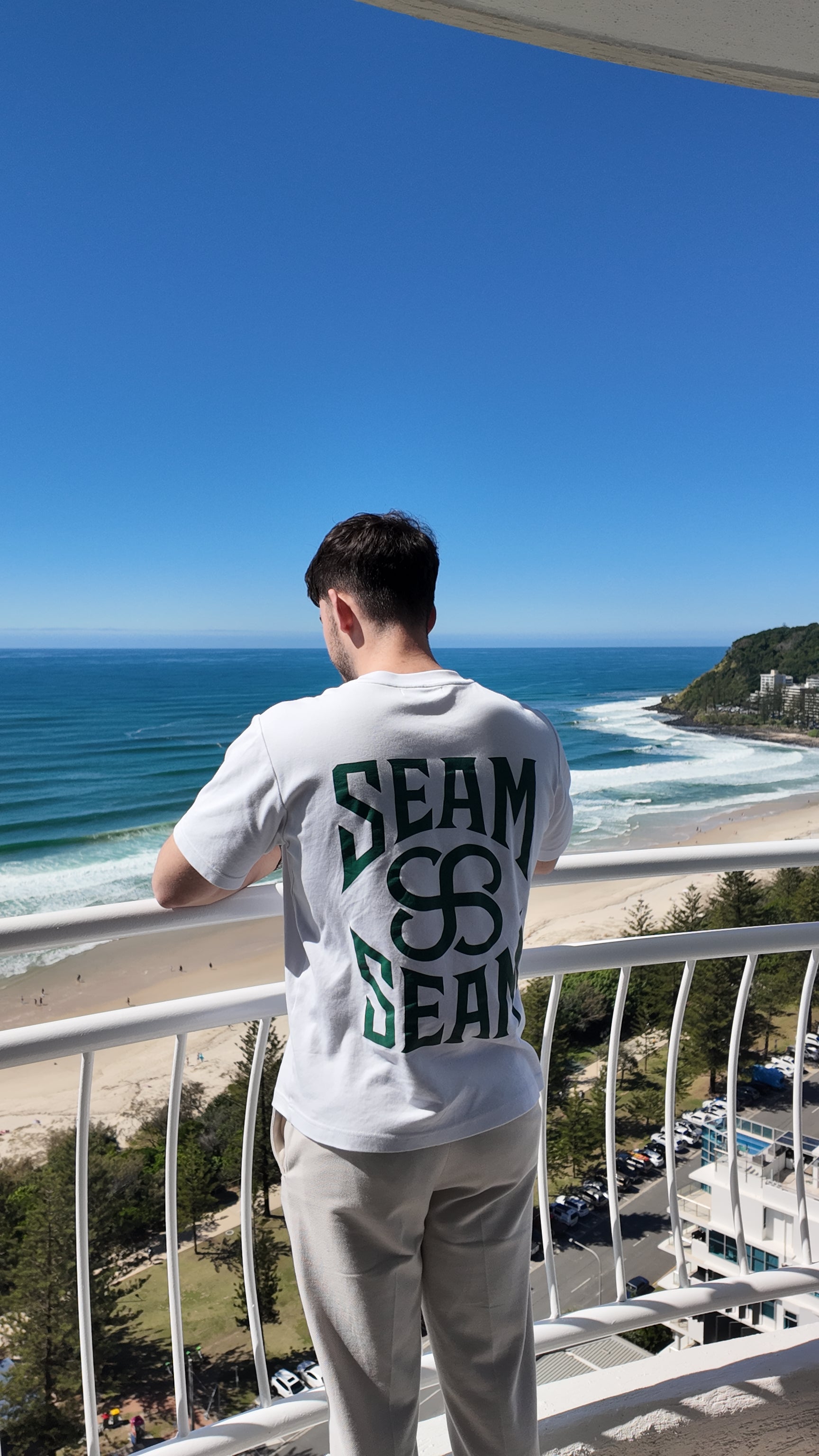Person wearing a white t-shirt with 'SEAM' printed on it, standing on a balcony overlooking the ocean.