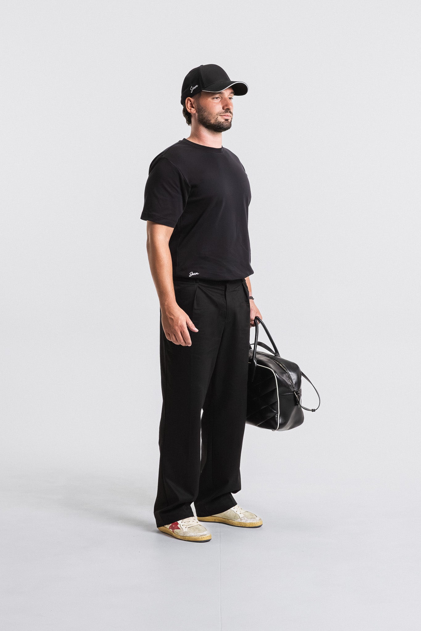 Man wearing a black t-shirt, pants, and cap, holding a black bag on a white background