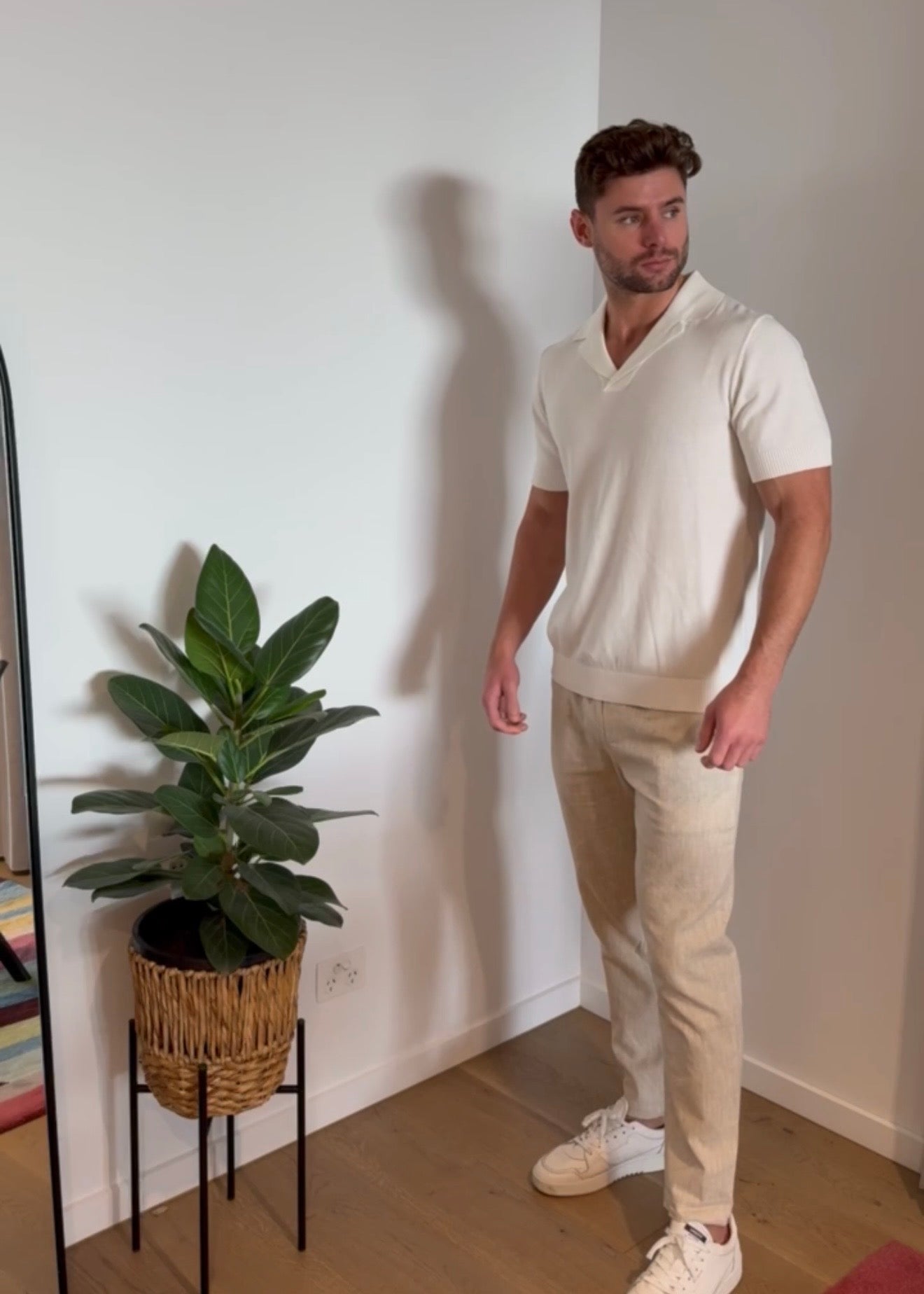 Man wearing a white shirt and beige pants standing next to a plant indoors.