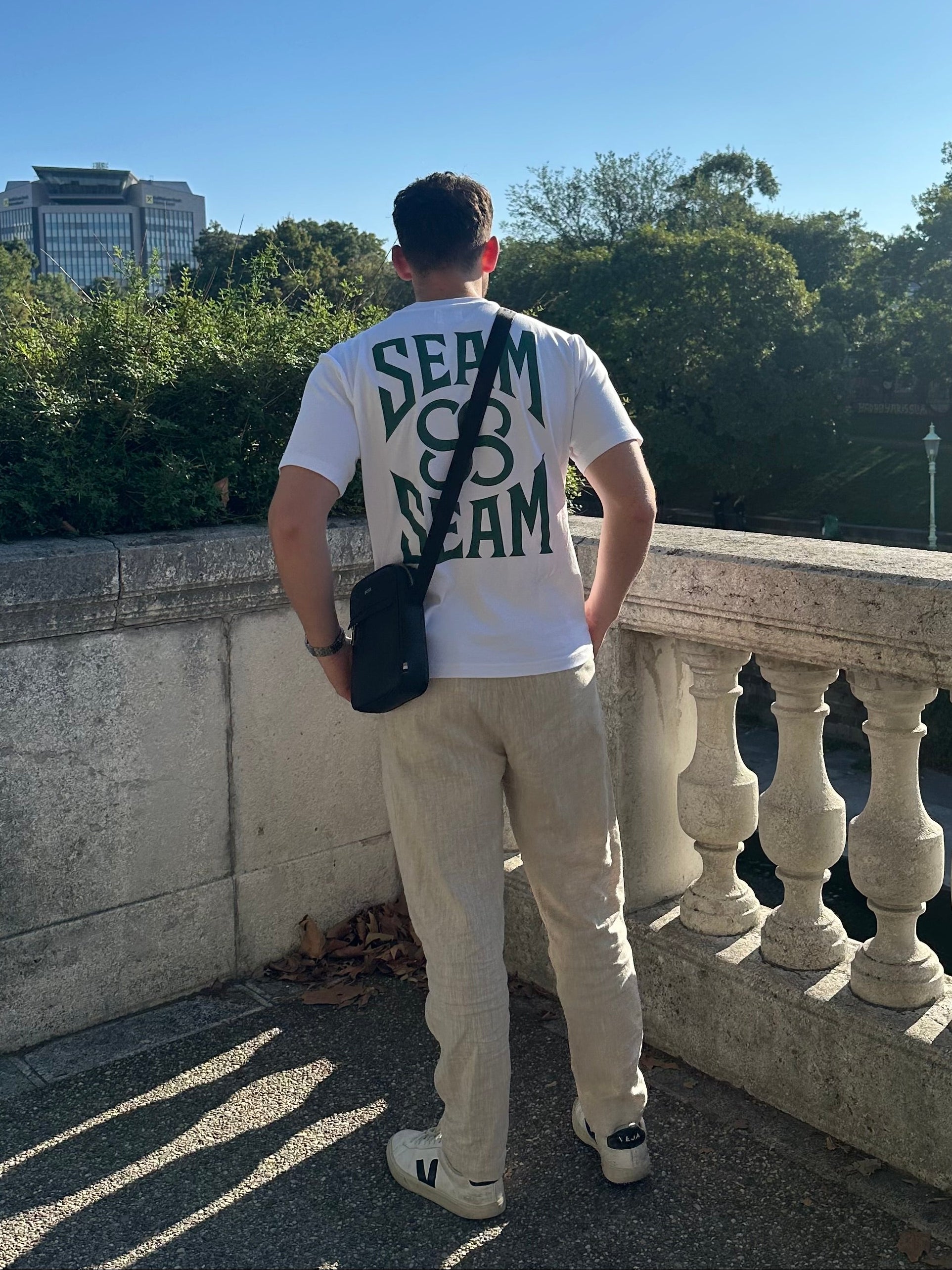 Person wearing a white t-shirt with text on the back, standing on a balcony with trees and buildings in the background.